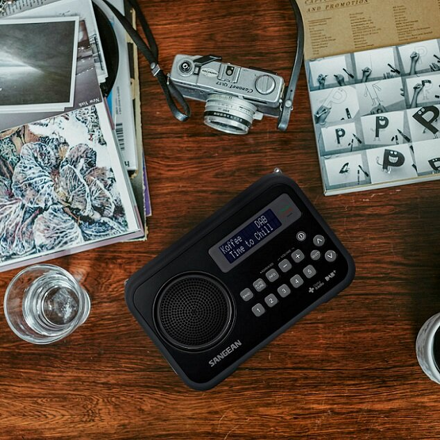 Czarne radio DAB na drewnianym stole, z aparatem i czasopismami. Ekran pokazuje 'Time to Chill'.