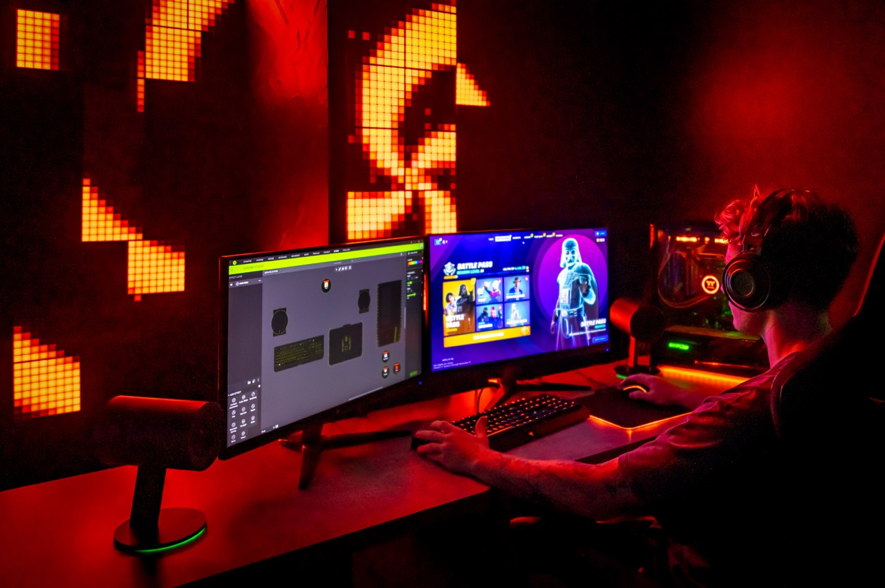 Una persona gioca su due monitor in una stanza buia illuminata da luci rosse. Indossano delle cuffie.