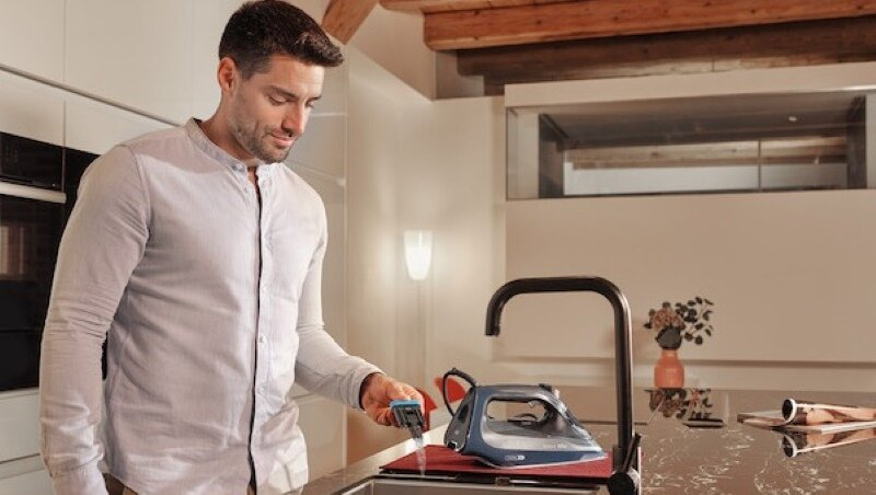 Mężczyzna w białej koszuli prasuje ubrania w pobliżu zlewu kuchennego.