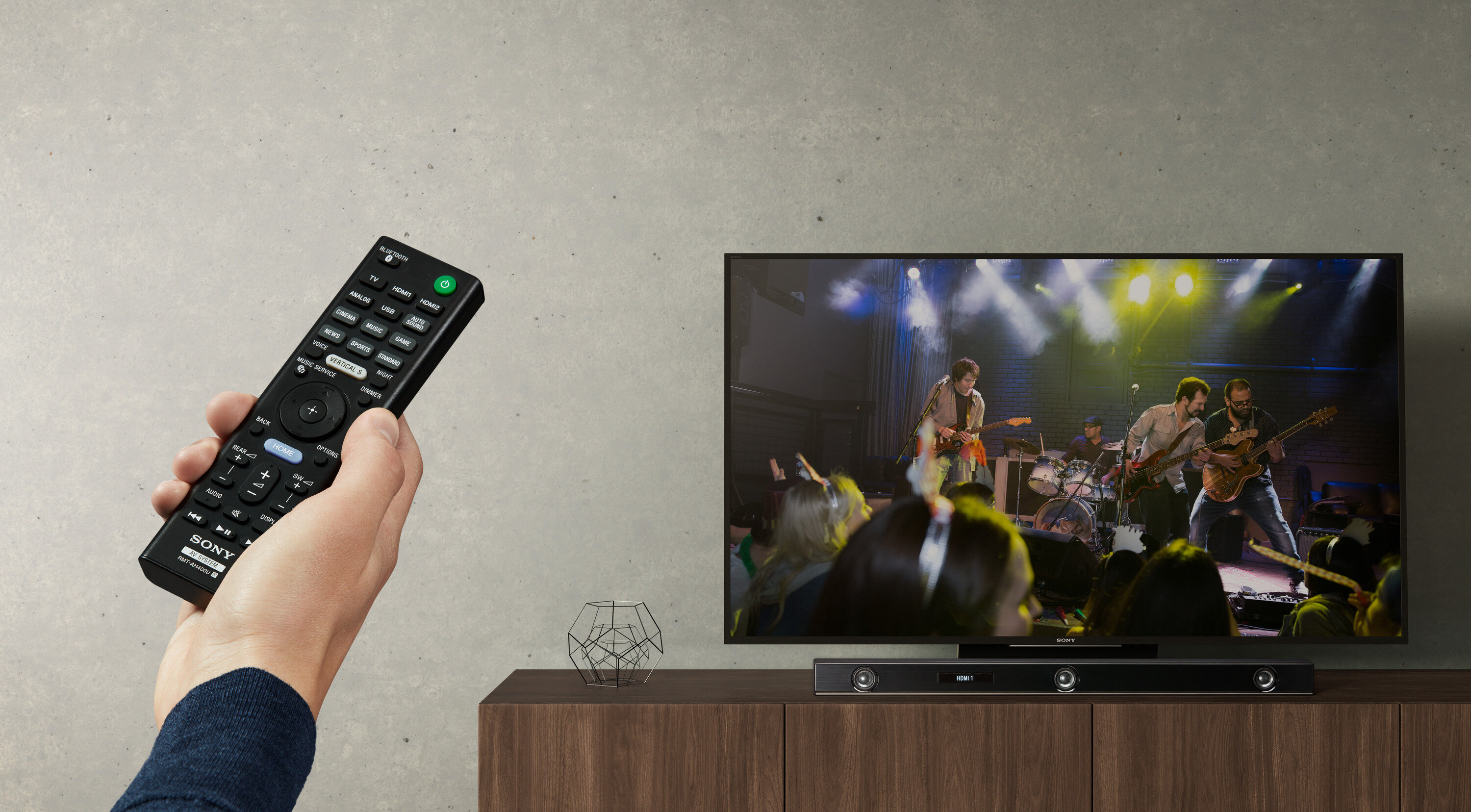 Osoba trzymająca pilota, oglądająca zespół grający w telewizji, z soundbarem na szafce poniżej.