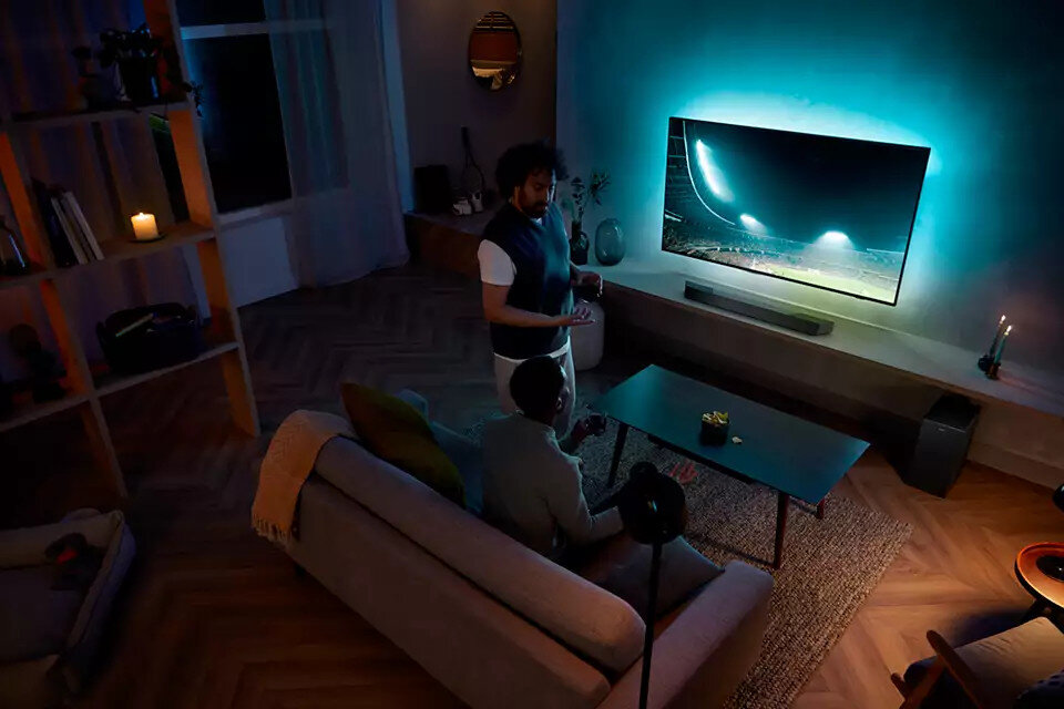 Salon z telewizorem pokazującym stadion, oświetlony niebieskim i białym światłem. Obecne dwie osoby.