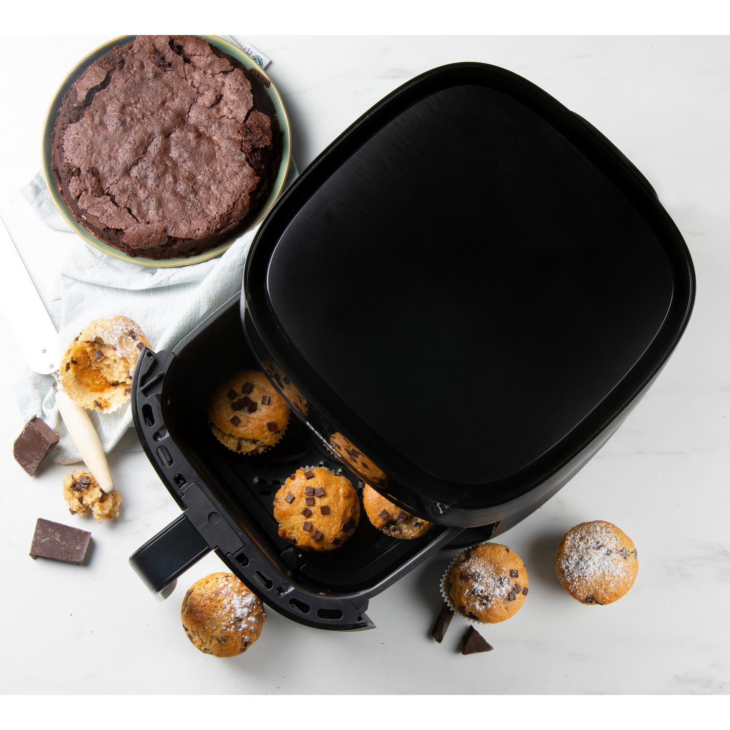 Une friteuse à air avec des muffins à l'intérieur et un brownie sur le côté, avec des ingrédients sur une surface en marbre.