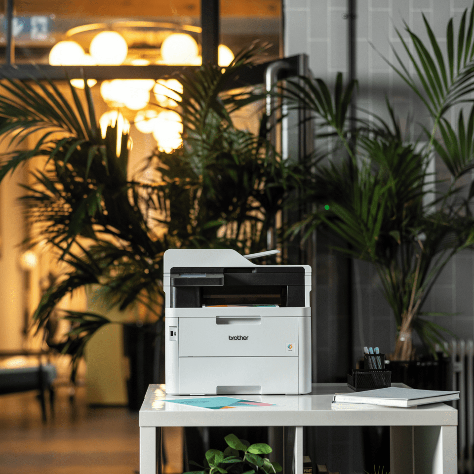 Una impresora Brother blanca sobre una mesa blanca con un fondo de plantas y una ventana.