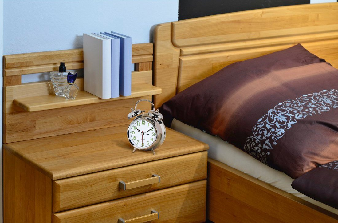 Drewniana szafka nocna z łóżkiem, książkami i zegarem. Łóżko ma brązową kołdrę i poduszki.