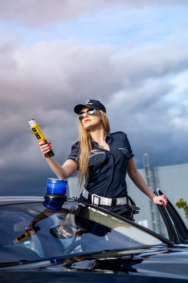 Policjantka w mundurze trzyma urządzenie przy samochodzie pod zachmurzonym niebem. Nosi okulary przeciwsłoneczne.