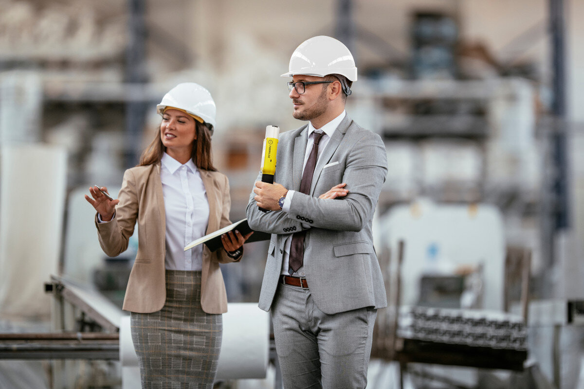 Dwie osoby w kaskach i garniturach patrzą w bok w otoczeniu przemysłowym.