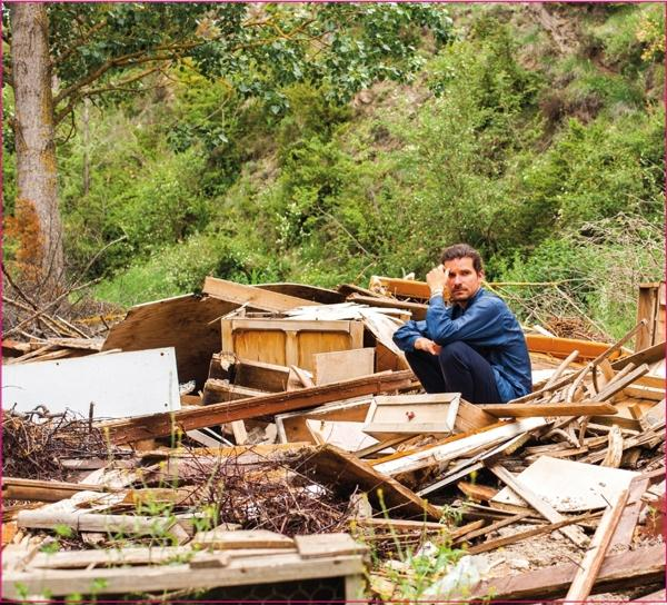 Ein Mann sitzt nachdenklich auf einem Holzhaufen im Freien, in einer natürlichen Umgebung.