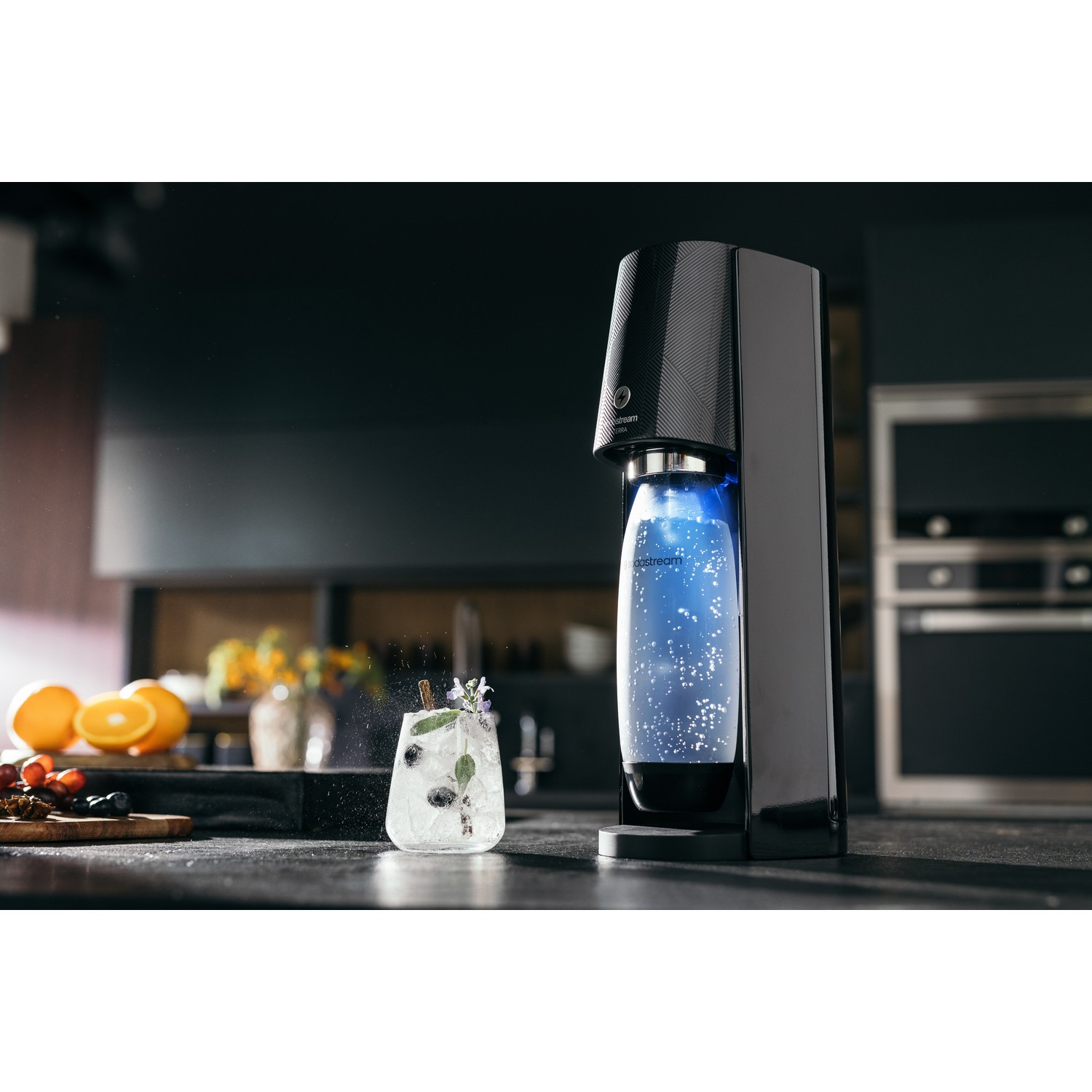 A soda machine with a bottle filling with blue liquid, a cocktail, and oranges on a counter.