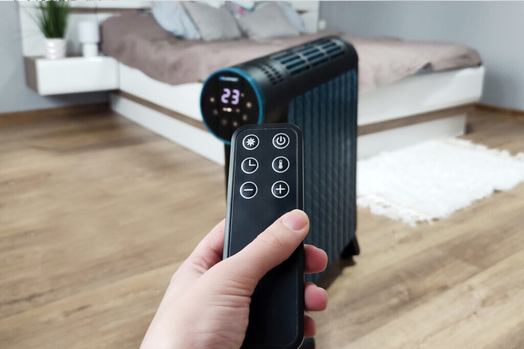Dłoń trzyma czarny pilot przed grzejnikiem i łóżkiem.