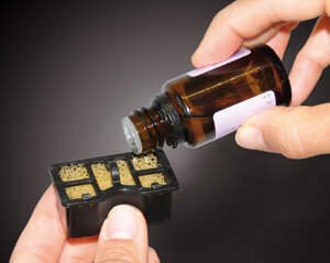 Dłonie wlewające płyn z butelki do czarnego pojemnika.