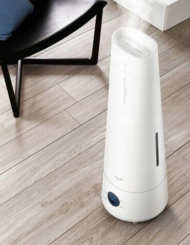 Biały nawilżacz na drewnianej podłodze, obok czarnego krzesła. Para wydobywa się z góry. Krzesło ma niebieską poduszkę.