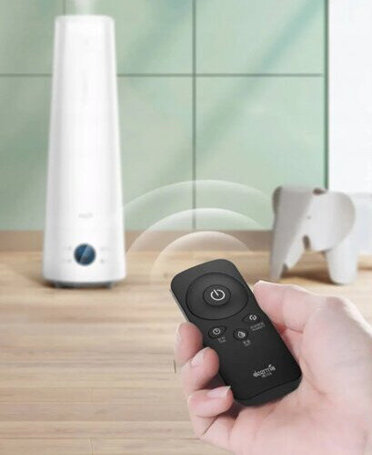 Ręka trzyma czarny pilot w pobliżu białego nawilżacza powietrza. W tle dekoracja w kształcie słonia.