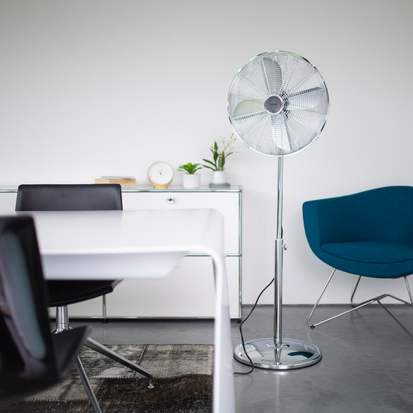 Srebrny wentylator w nowoczesnym biurze z białymi ścianami, biurkiem i turkusowym krzesłem.
