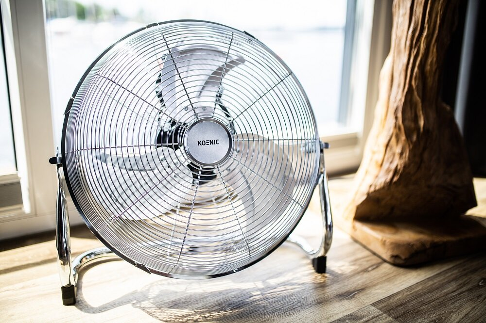 Srebrny wentylator podłogowy stoi na drewnianej podłodze przed oknem. Po prawej znajduje się drewniana rzeźba.