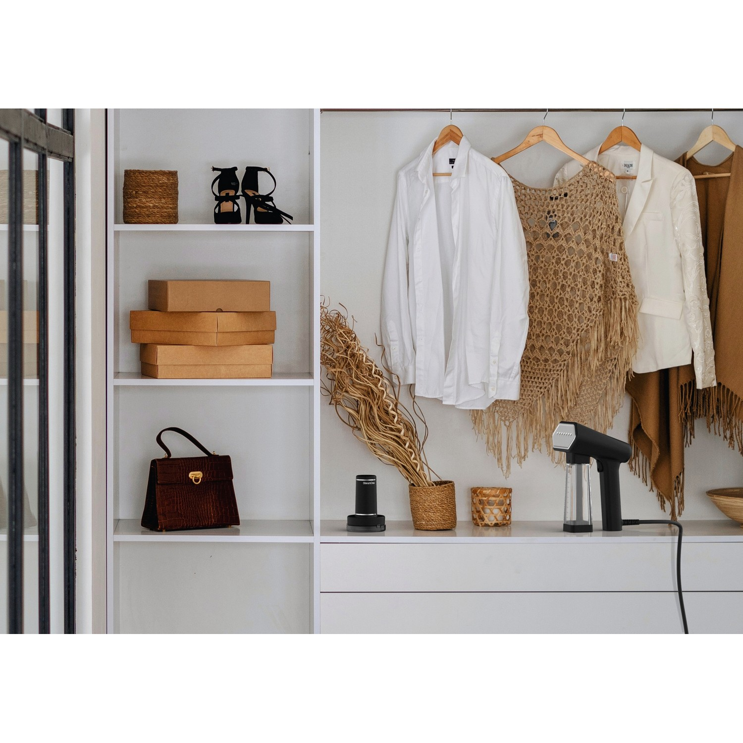Placard avec des vêtements suspendus, des boîtes et un défroisseur. Un sac à main noir et des chaussures sont visibles.