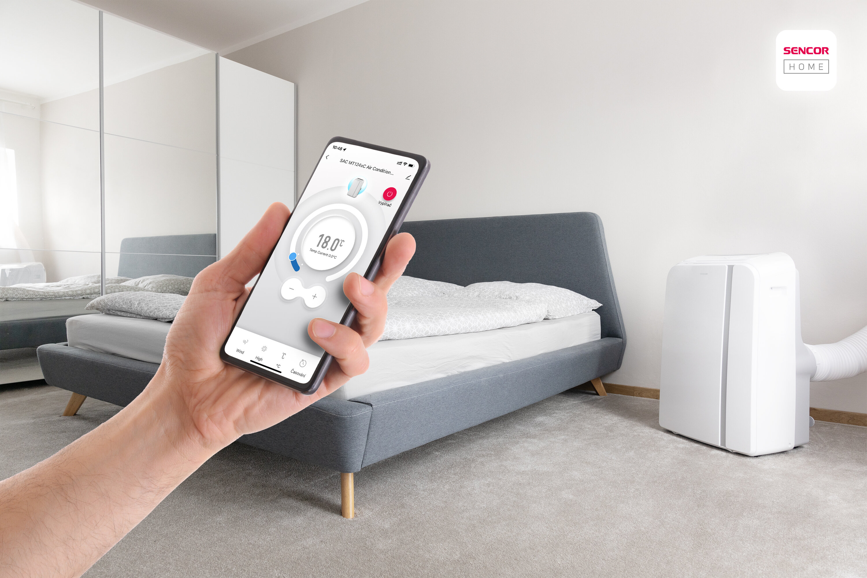 Ręka trzyma telefon sterujący klimatyzacją. Sypialnia z łóżkiem i białą jednostką klimatyzacji w tle.