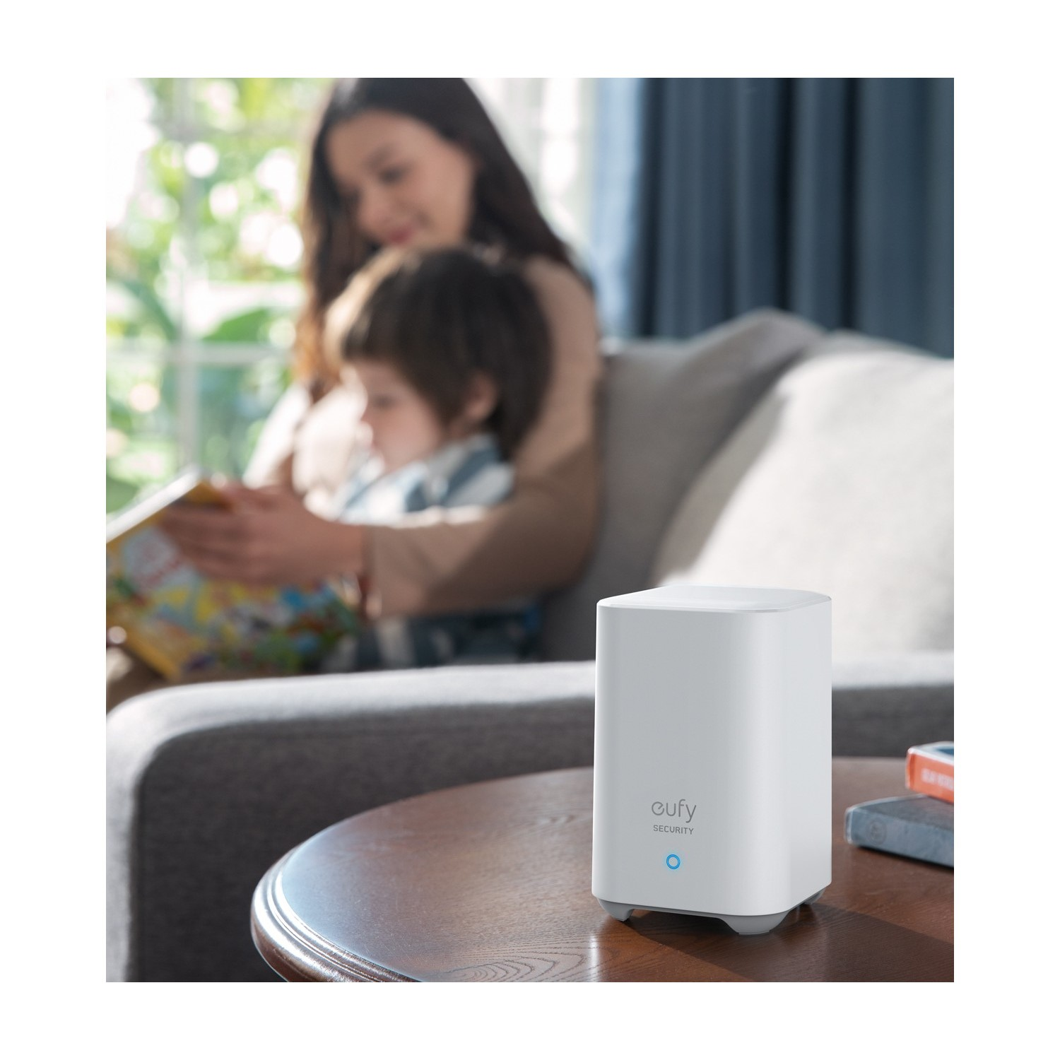 Un appareil domestique intelligent blanc sur une table, avec une femme et un enfant en arrière-plan lisant un livre.