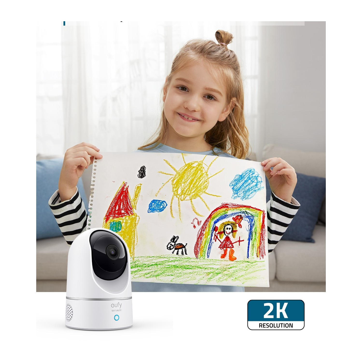 Une fille tient un dessin de maison, soleil et arc-en-ciel. Une caméra blanche au premier plan.