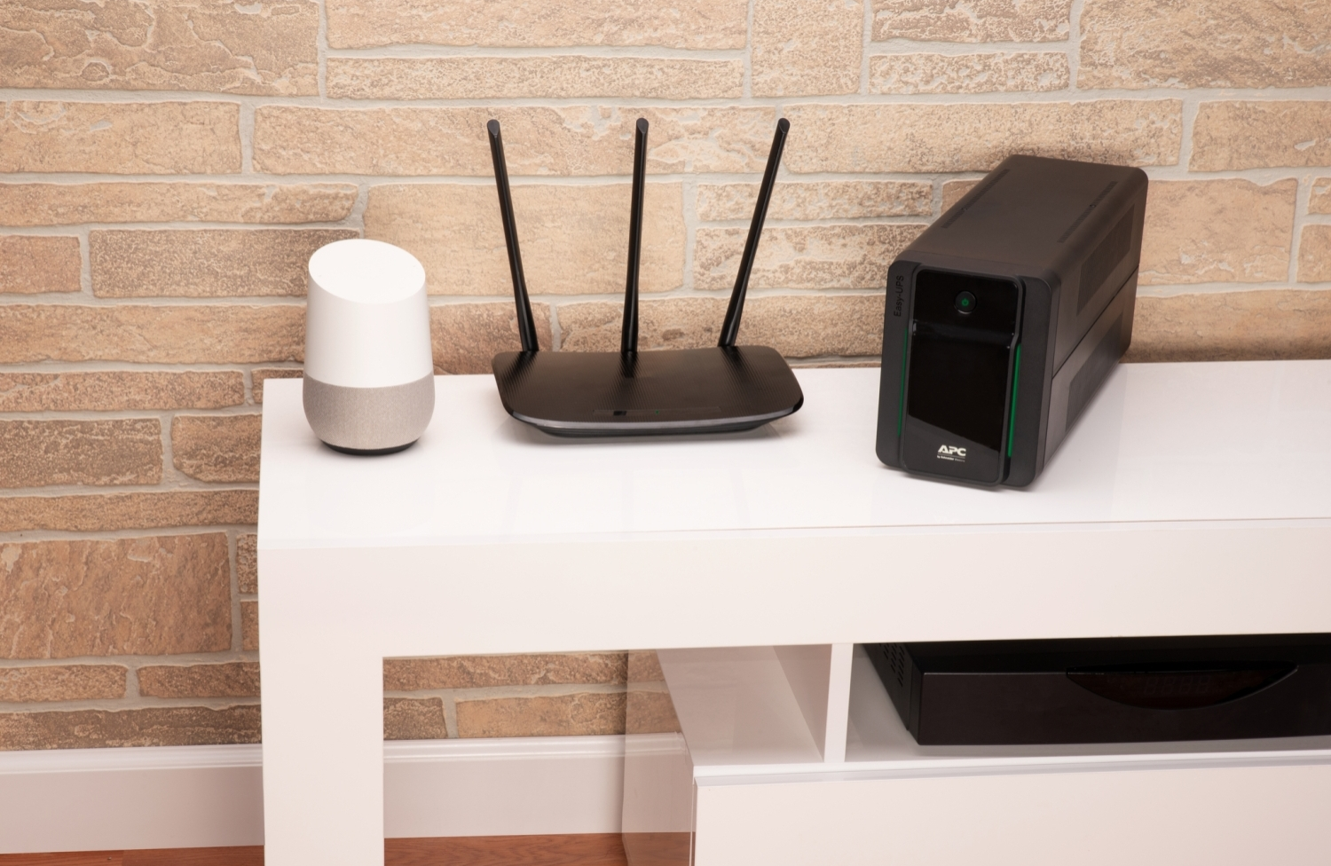 Egy fehér állványon Google Home, router és fekete szünetmentes tápegység van, téglaburkolat mellett.