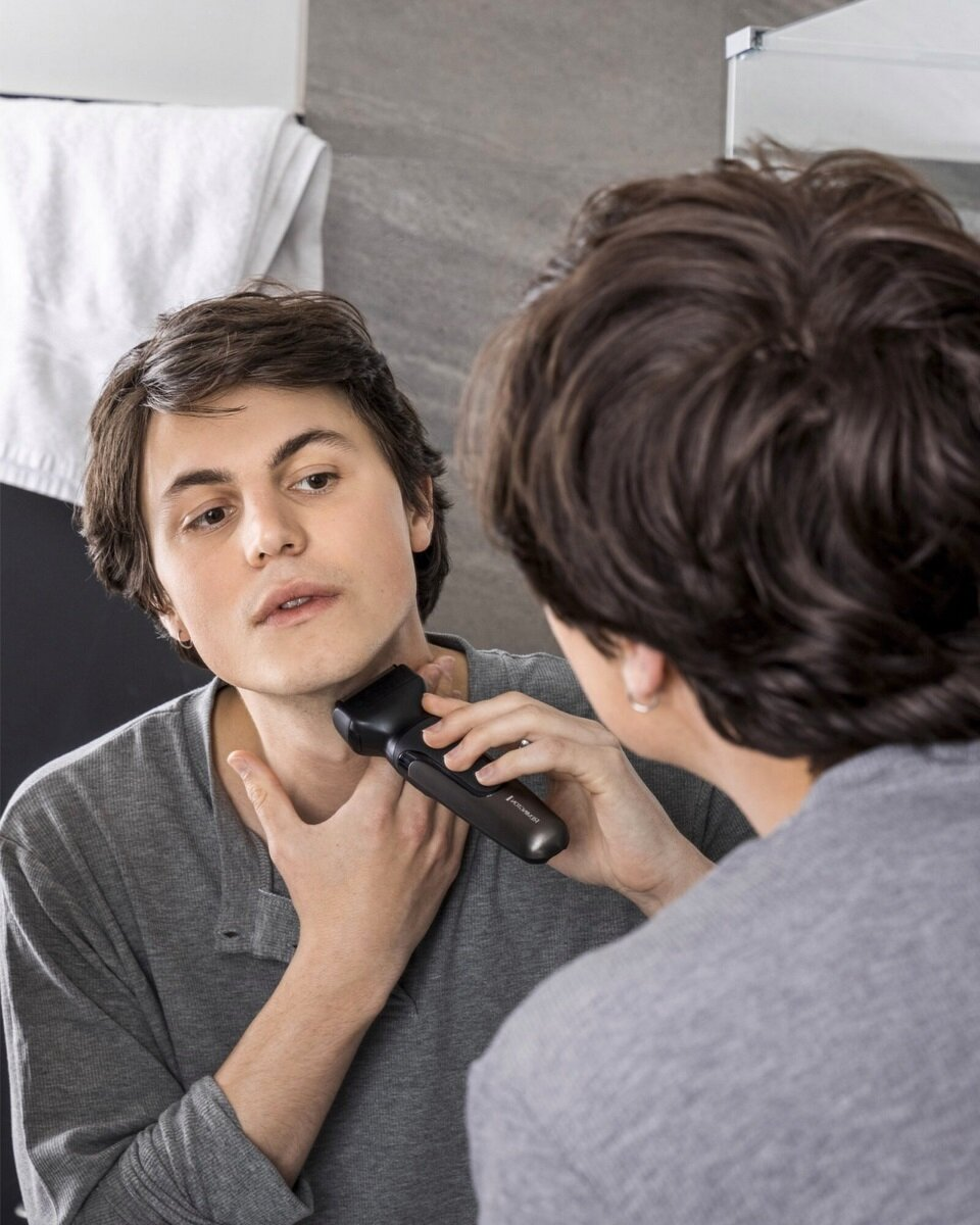 Mężczyzna goli szyję przed lustrem czarną golarką.