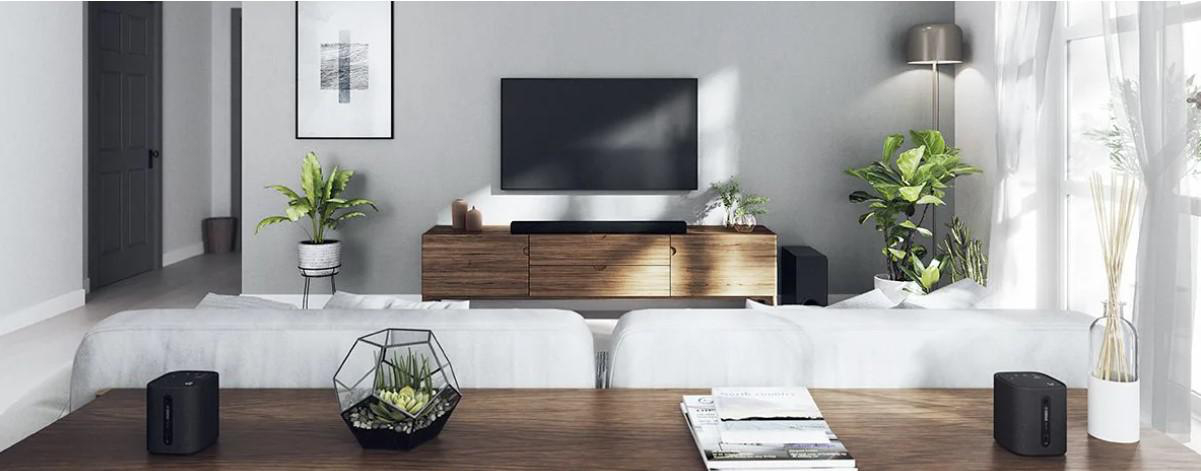 Ein Wohnzimmer mit Sofa, Fernseher auf einem Holzschrank und Soundbar. Zwei Lautsprecher und Pflanzen sind vorhanden.