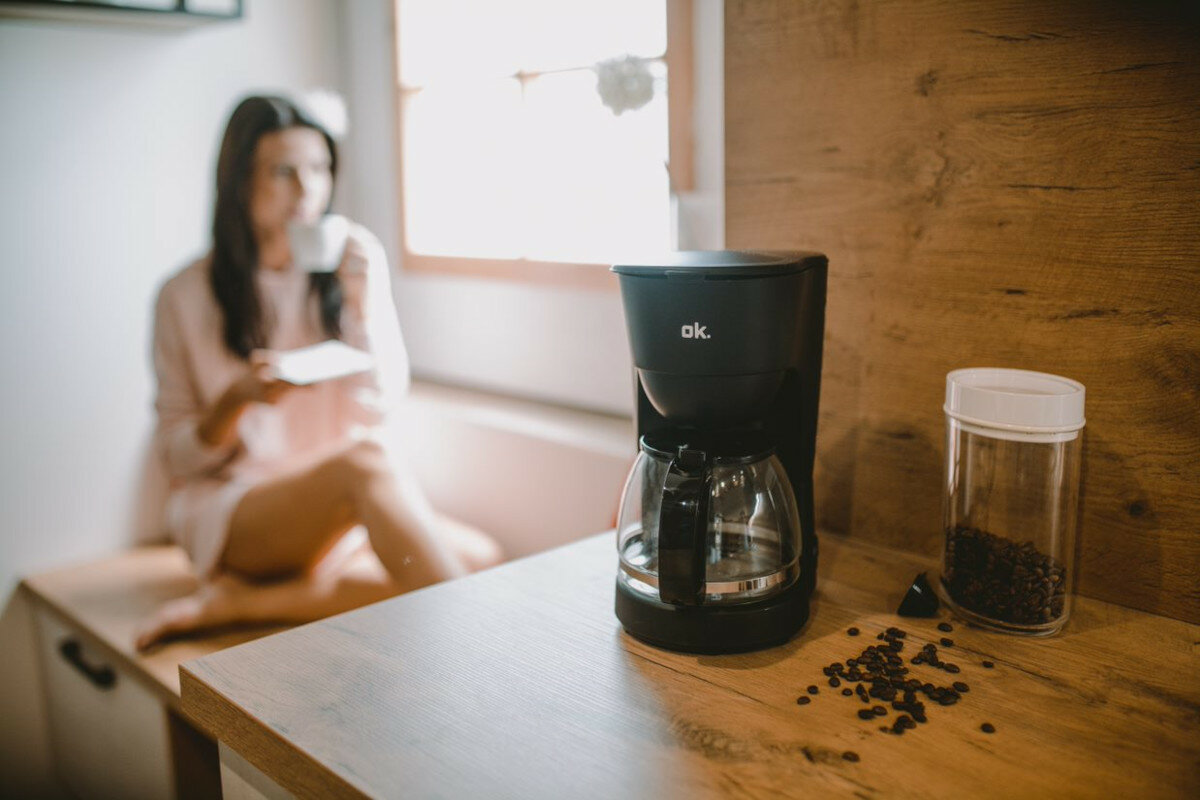 Czarny ekspres do kawy ze szklaną karafką i ziarnami na drewnianym blacie. Kobieta w tle.