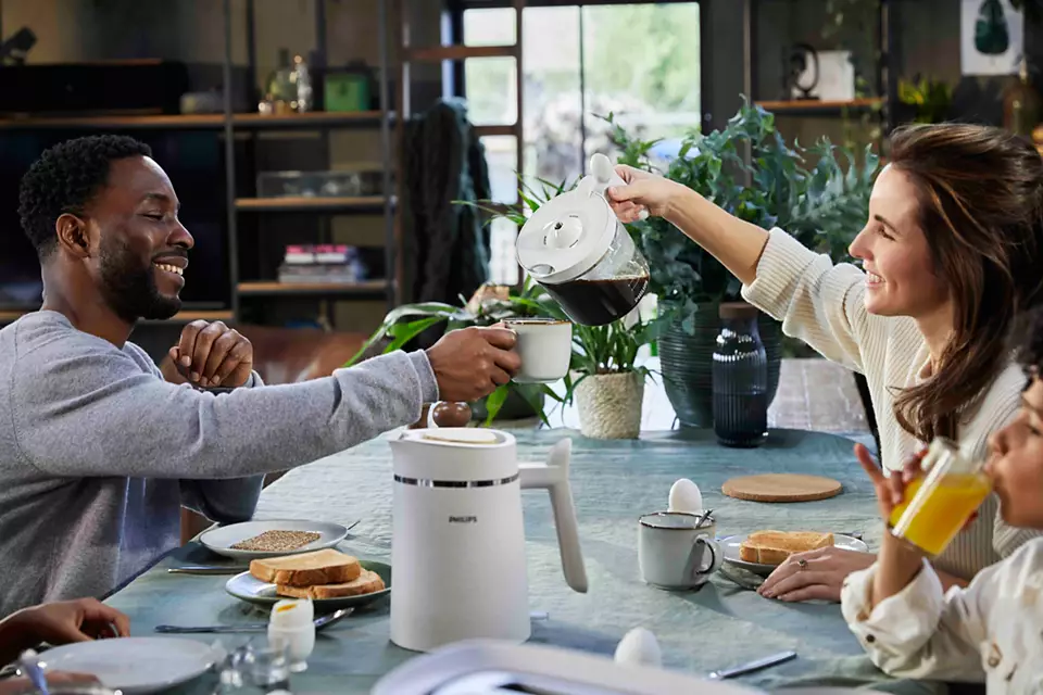 Rodzina je śniadanie. Kobieta nalewa kawę. Tosty, jajka i ekspres do kawy są na stole.