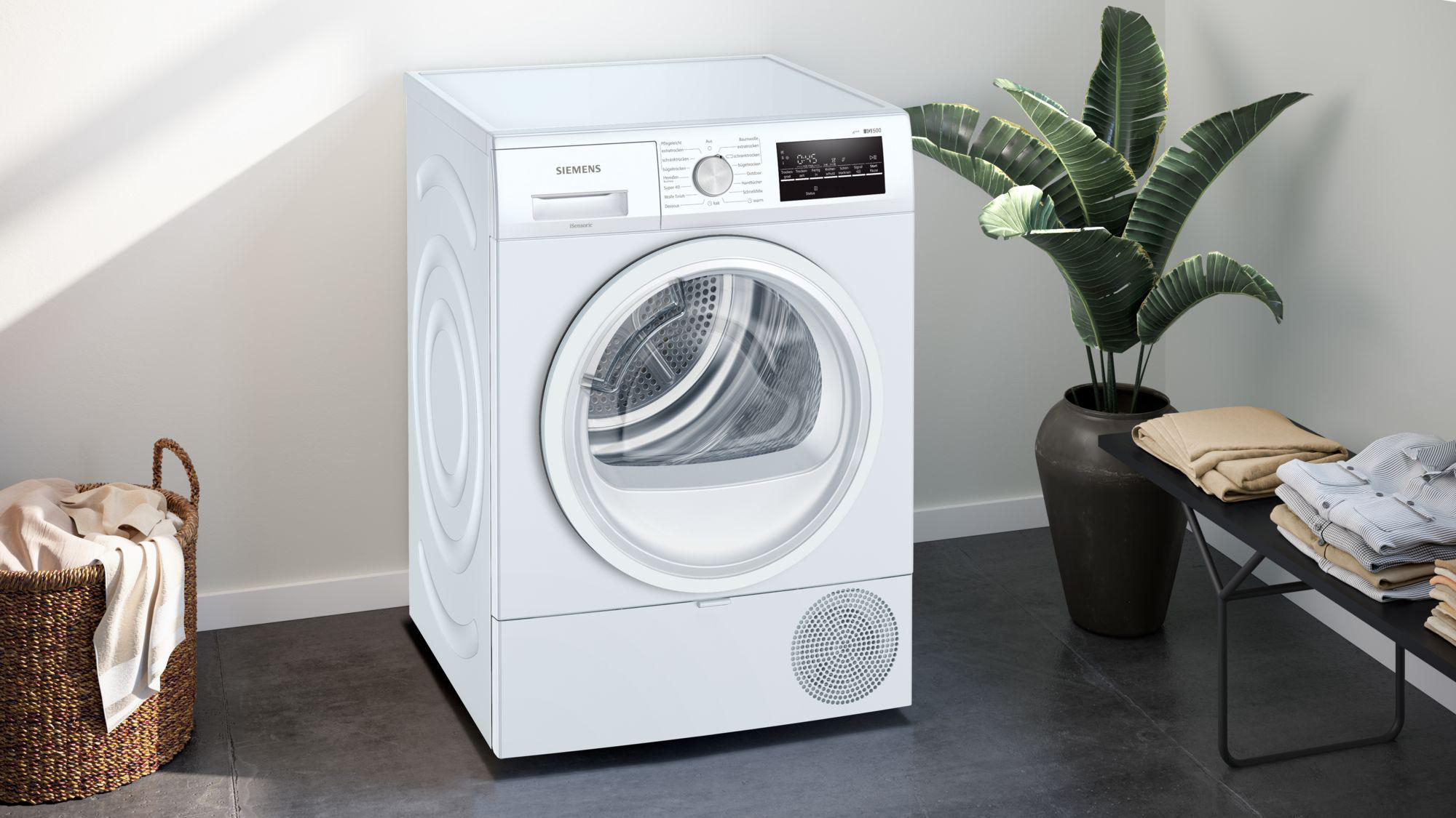 Ein weißer Siemens-Trockner in einem Raum. Ein Wäschekorb und eine Zimmerpflanze stehen neben dem Trockner.