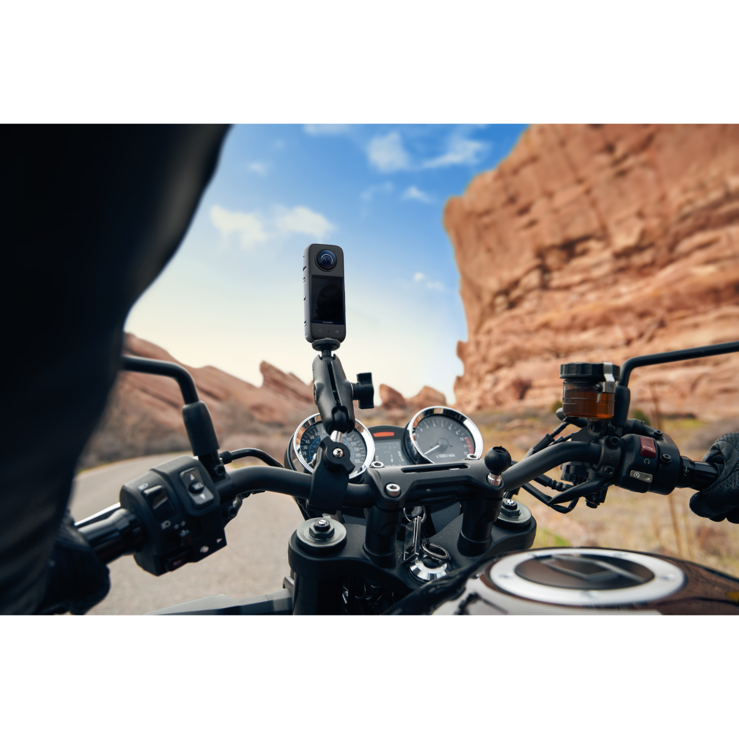 Motorcycle handlebars with a 360° camera mounted, against a red rock background.