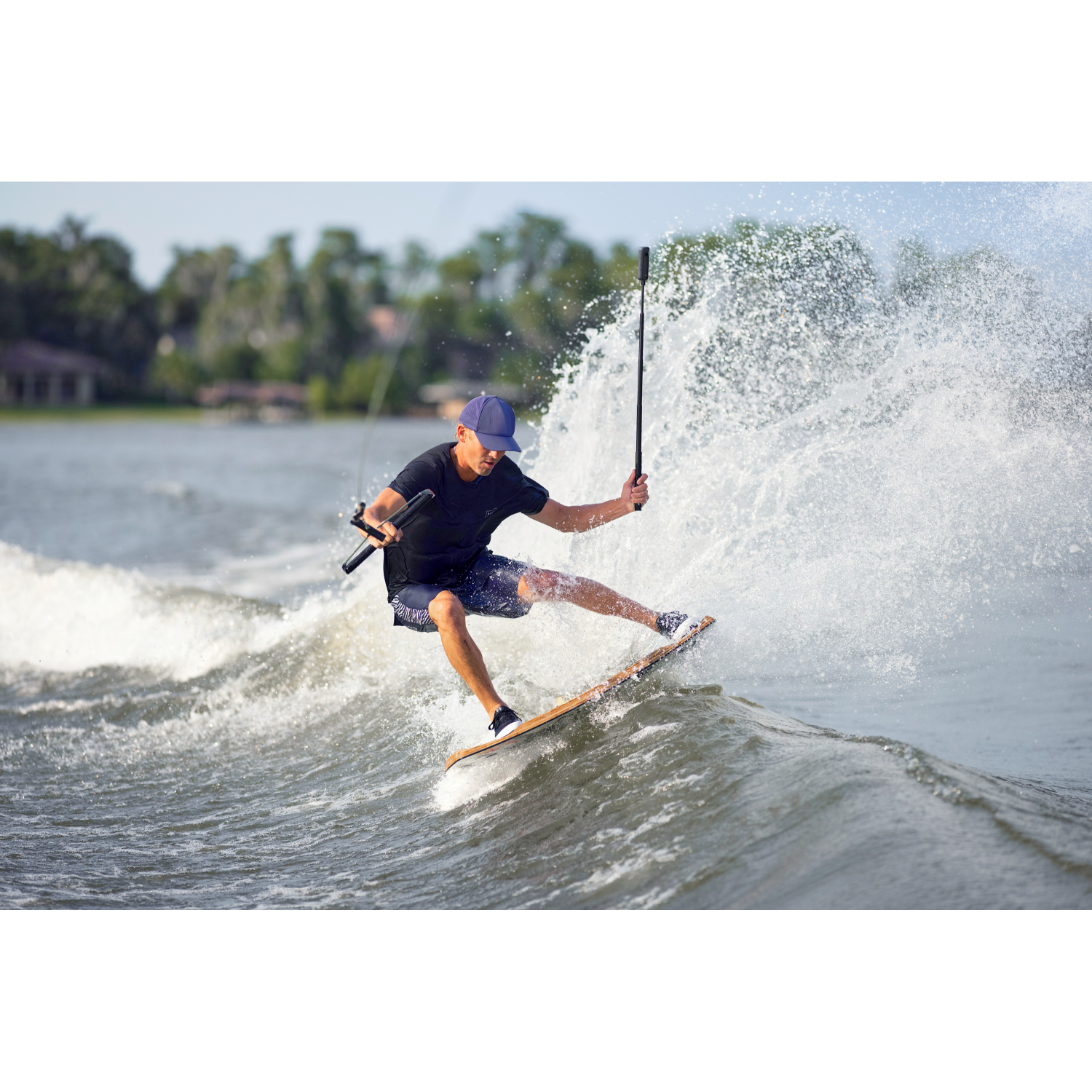 Man wakeboarding. He balances on board, with water splashing. Blue hat, black shirt.