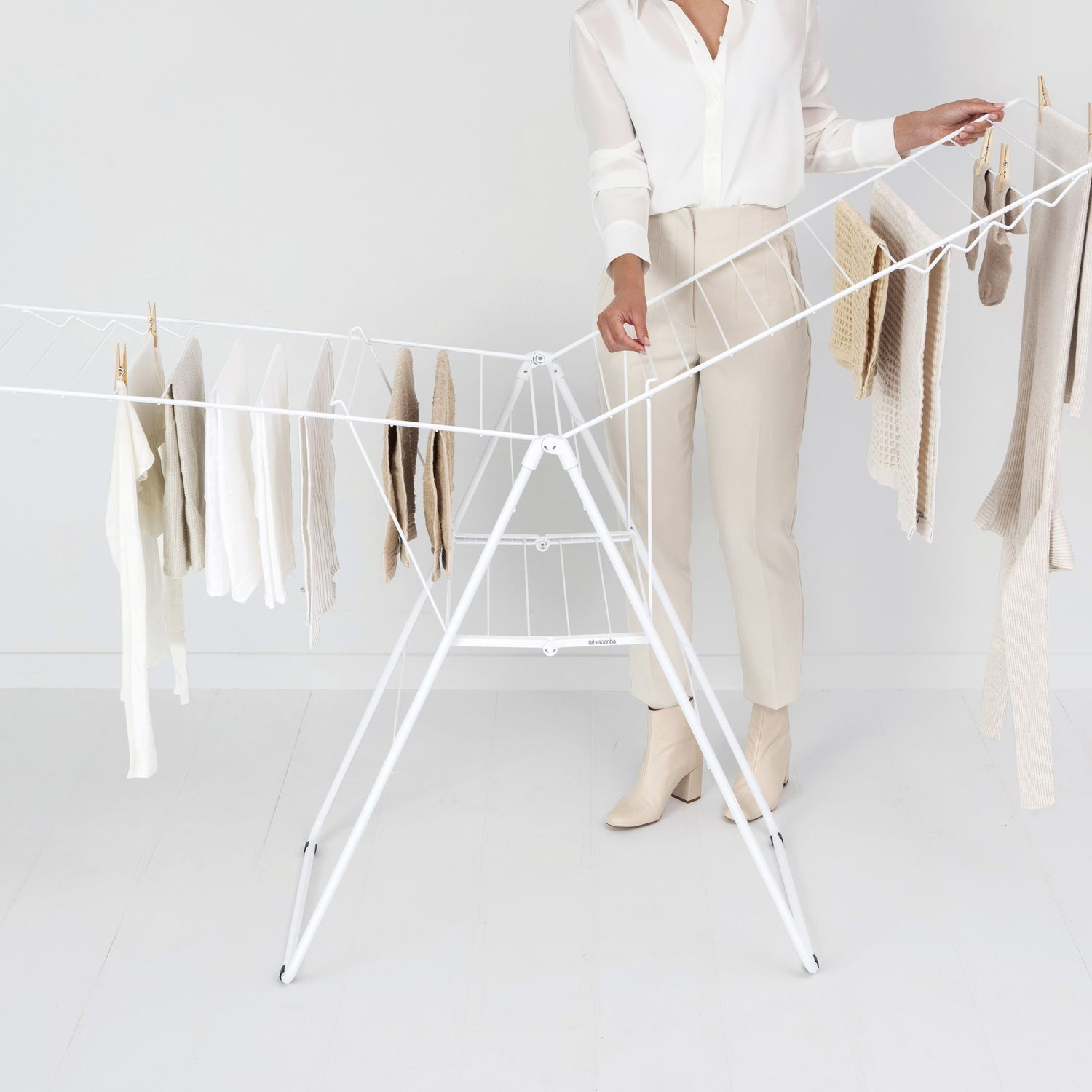 Vrouw hangt kleding op een witte droogrek. Kleding is wit en beige. Witte achtergrond.