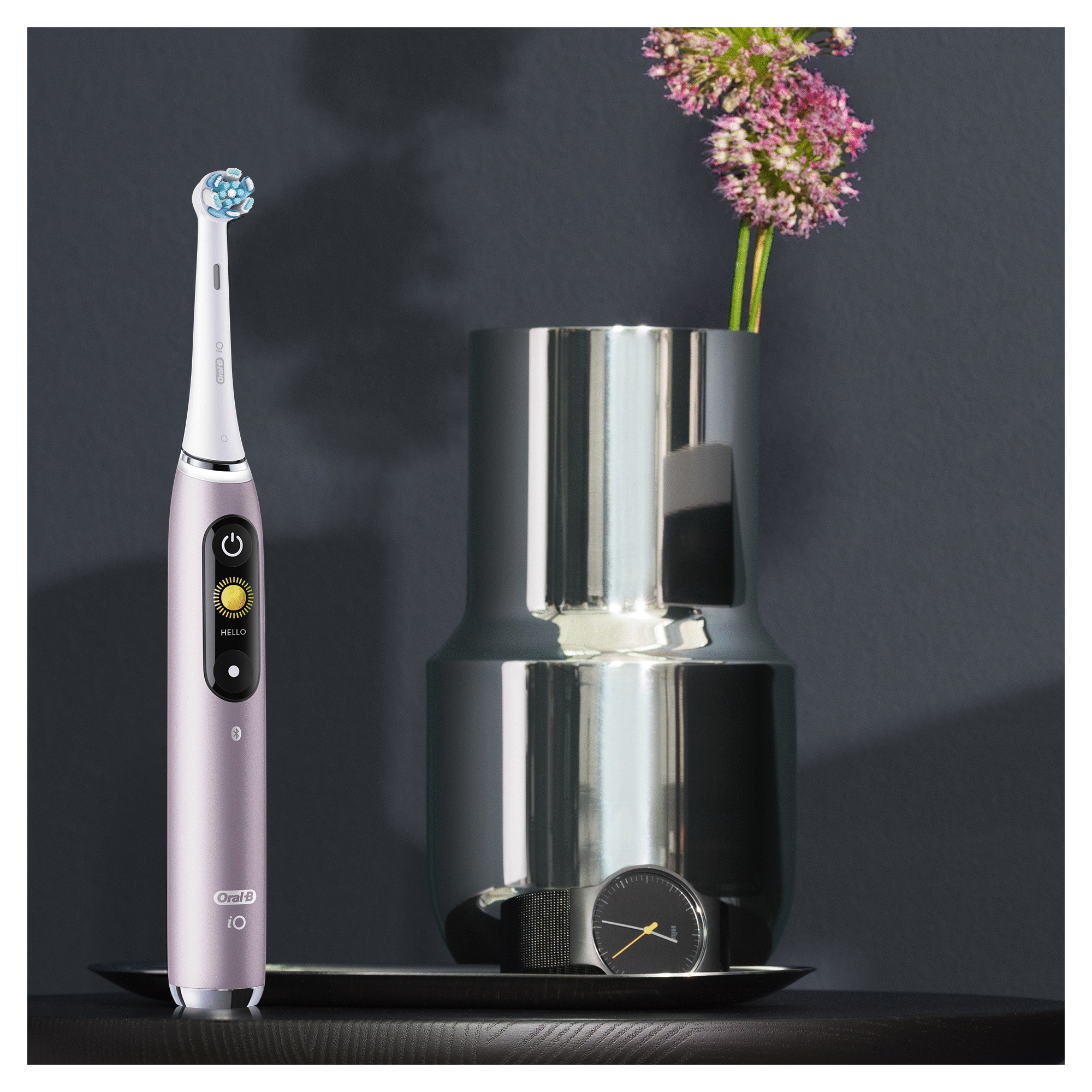 Une brosse à dents électrique et un vase avec des fleurs. La brosse est rose et le vase est argenté. Fond sombre.