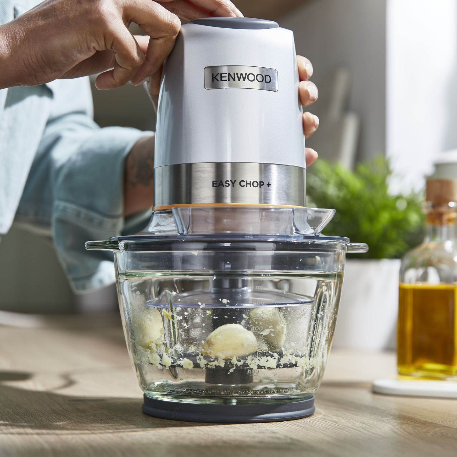 Een zilver- en witte food chopper met een doorzichtige kom, gevuld met knoflook. Handen zijn zichtbaar.