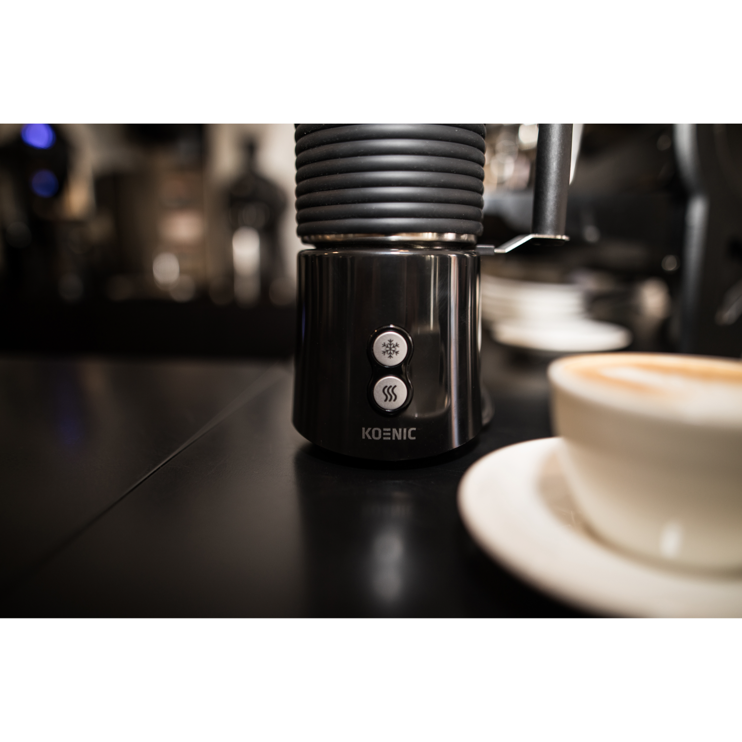 Black milk frother with two buttons and a cup of coffee on a black counter; blurred background.