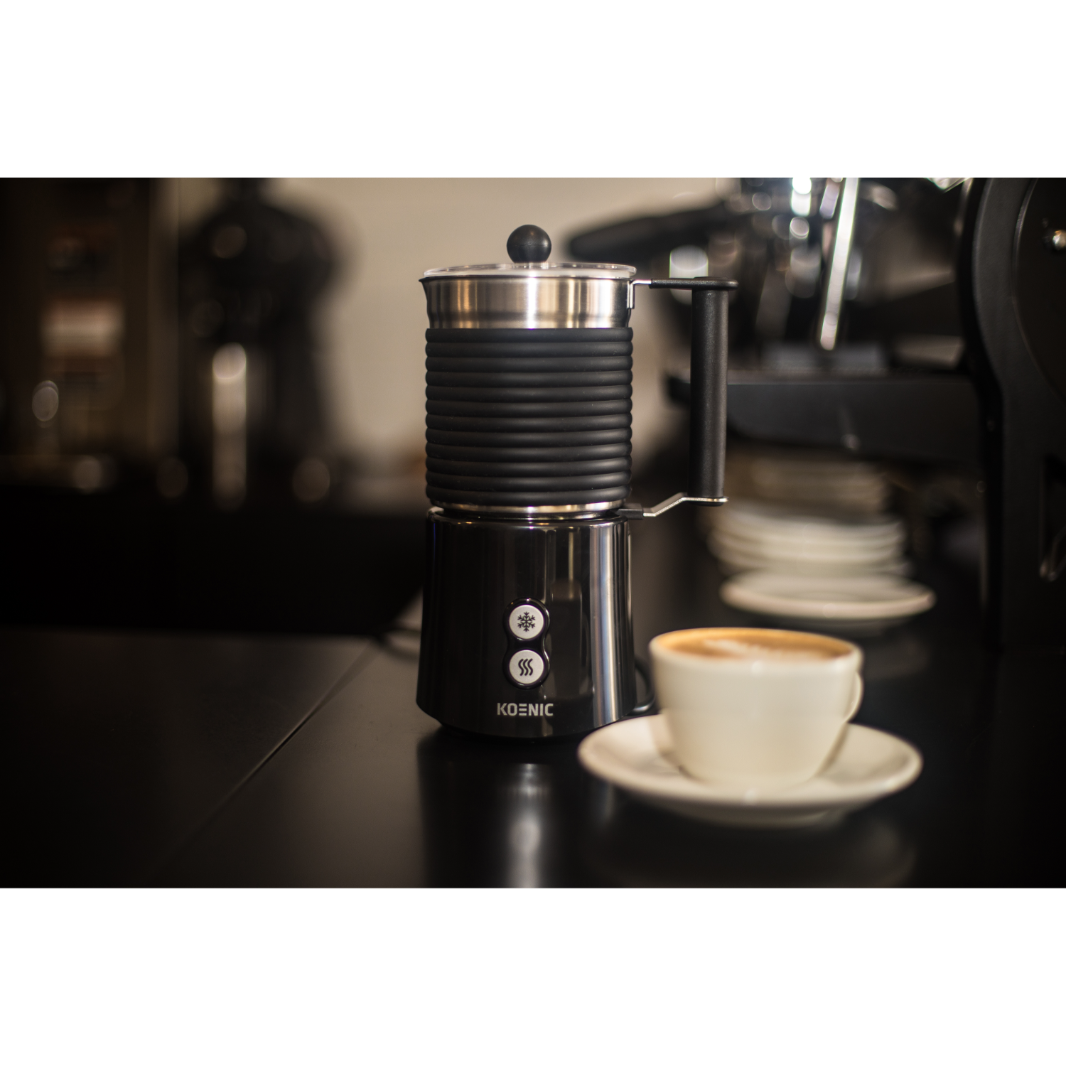 A black and silver milk frother with a cup of cappuccino on a black surface.
