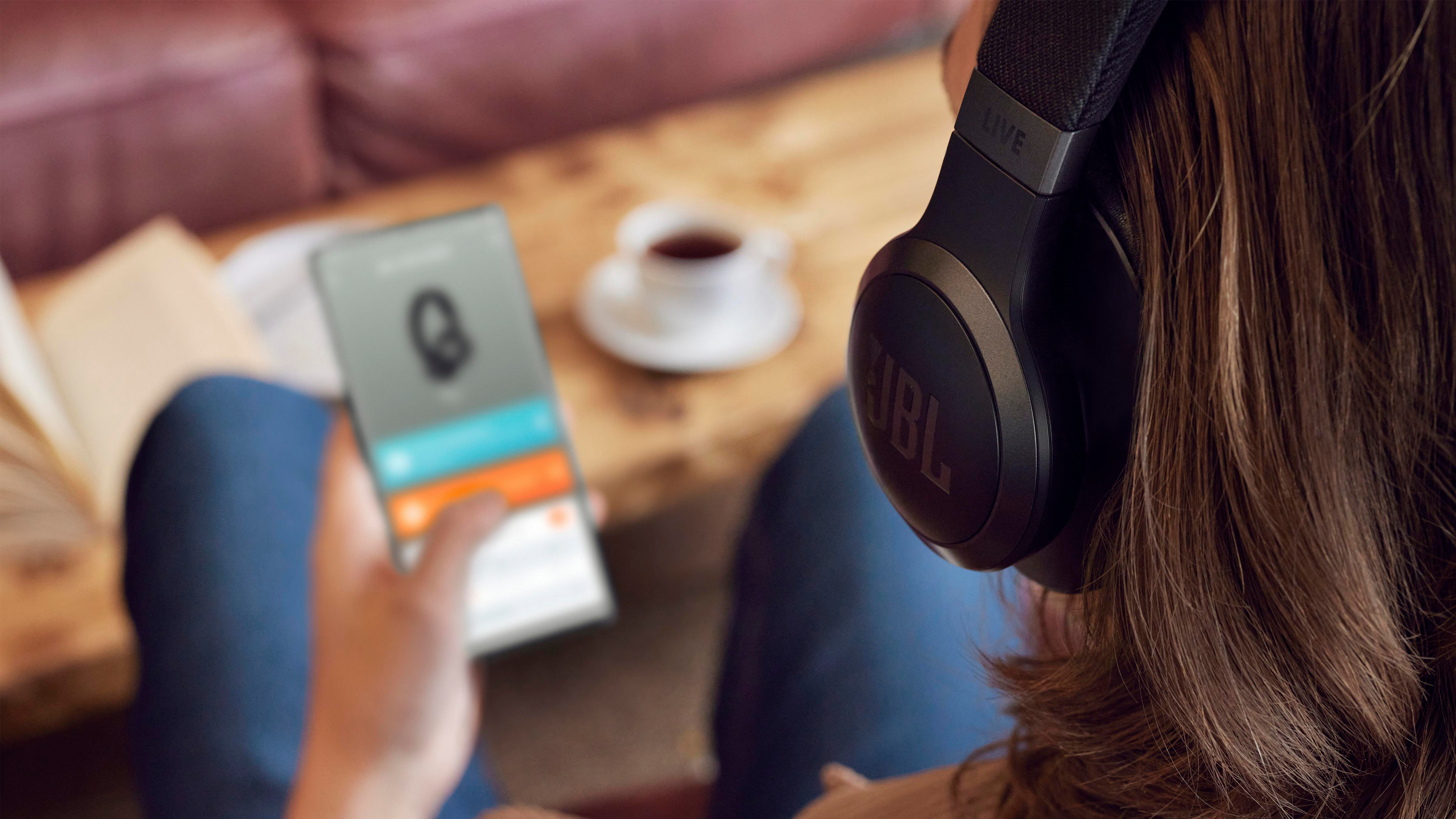Person mit schwarzen JBL-Kopfhörern, die ein Telefon benutzt. Eine Kaffeetasse steht auf dem Tisch.