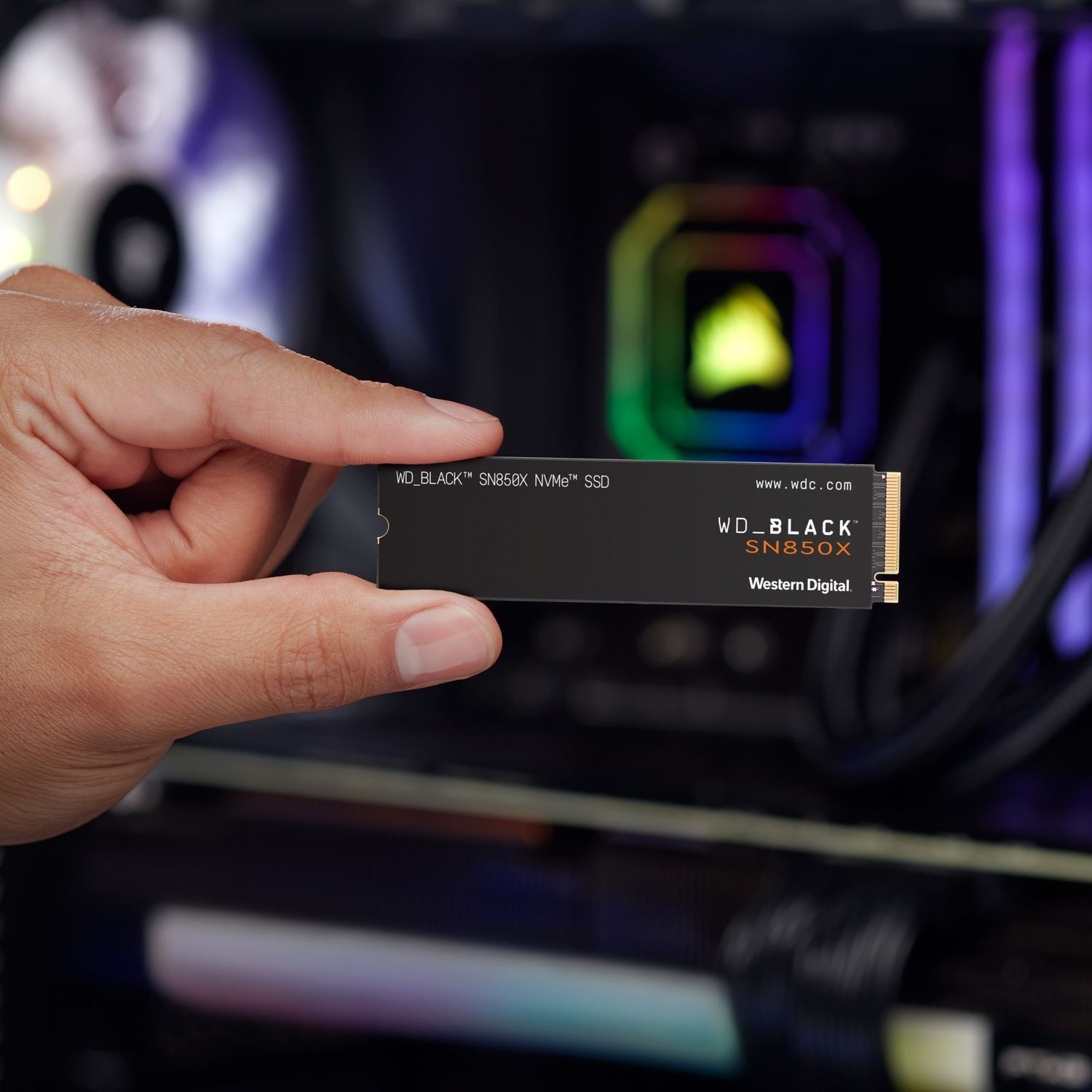 Een hand die een zwarte SSD vasthoudt, met een computer op de achtergrond.