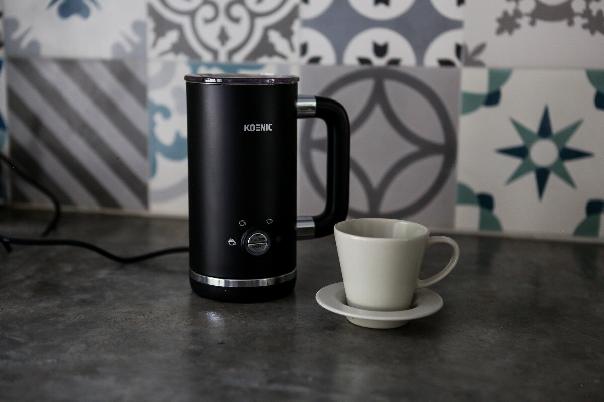 Czarny spieniacz do mleka z białą filiżanką i spodkiem. Ustawiony na szarym blacie.