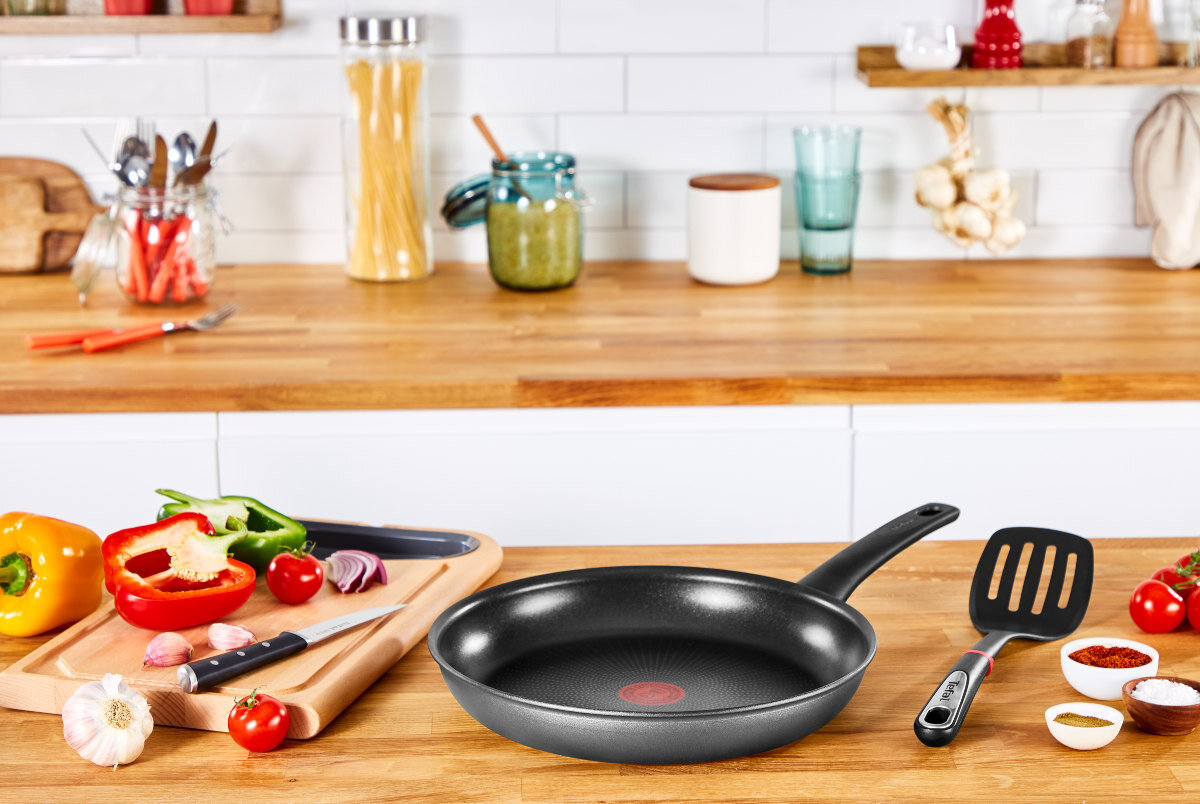 Patelnia, deska do krojenia z warzywami, nóż i przybory kuchenne na drewnianym blacie.