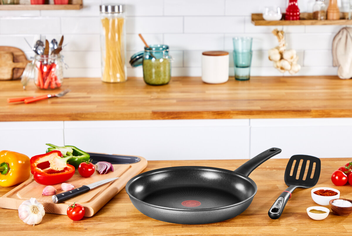 Scena kuchenna z patelnią, szpatułką, warzywami i przyprawami na drewnianym blacie.
