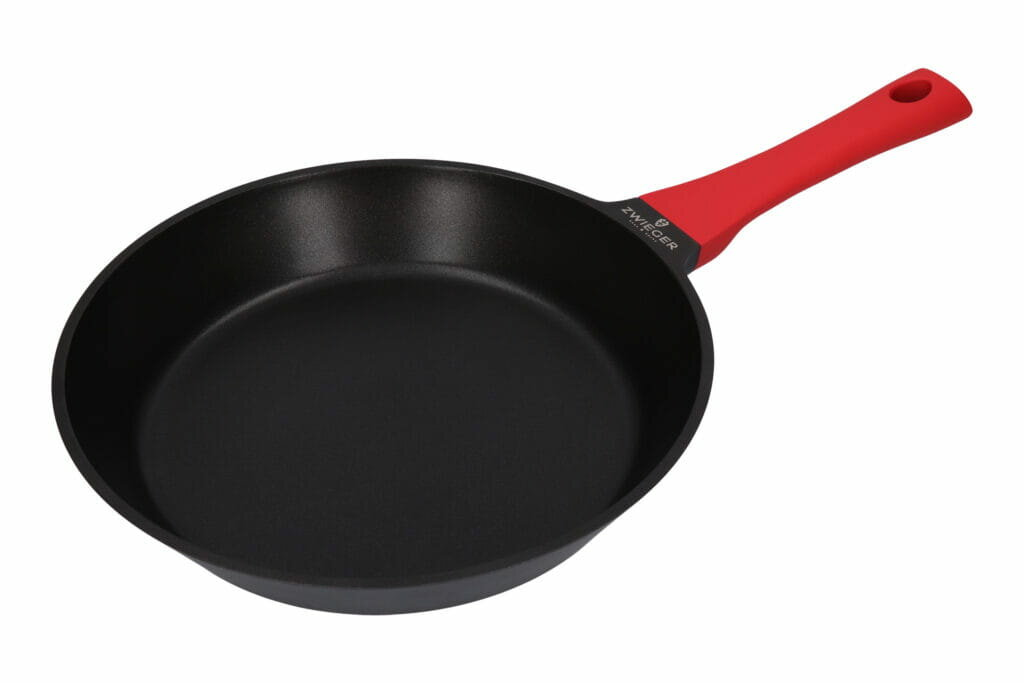 Czarna patelnia z czerwonym uchwytem. Uchwyt ma otwór na końcu. Marka jest na patelni.