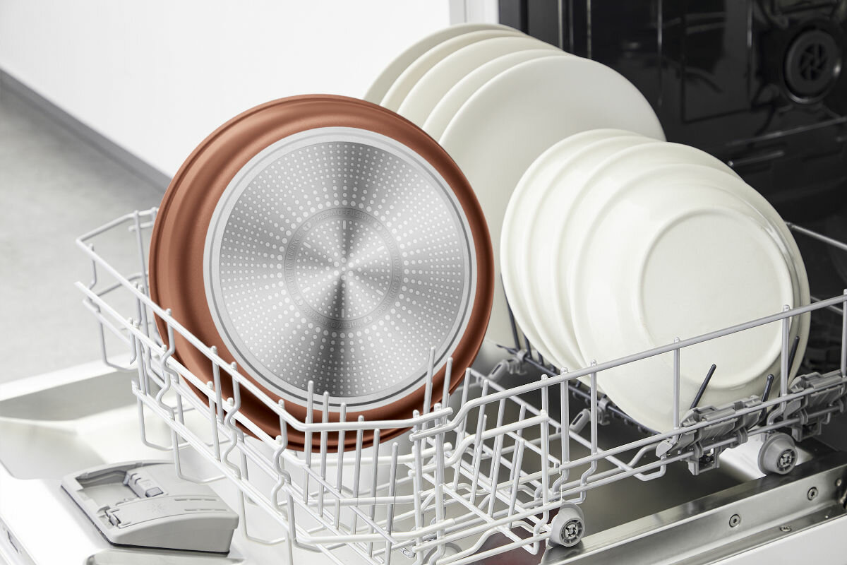 Zmywarka z patelnią o miedzianym dnie i białymi talerzami w środku.