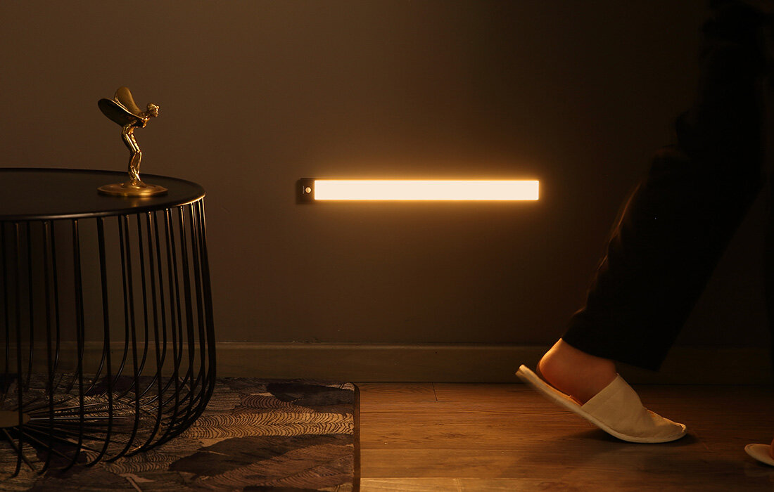 Stopa osoby w białym kapciu w pobliżu podświetlanego paska LED na ciemnej ścianie.