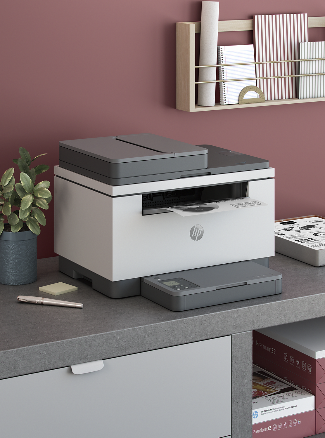 Une imprimante HP blanche et grise est sur un bureau gris. Une plante, un stylo et du papier sont devant. Des livres sont sur une étagère.