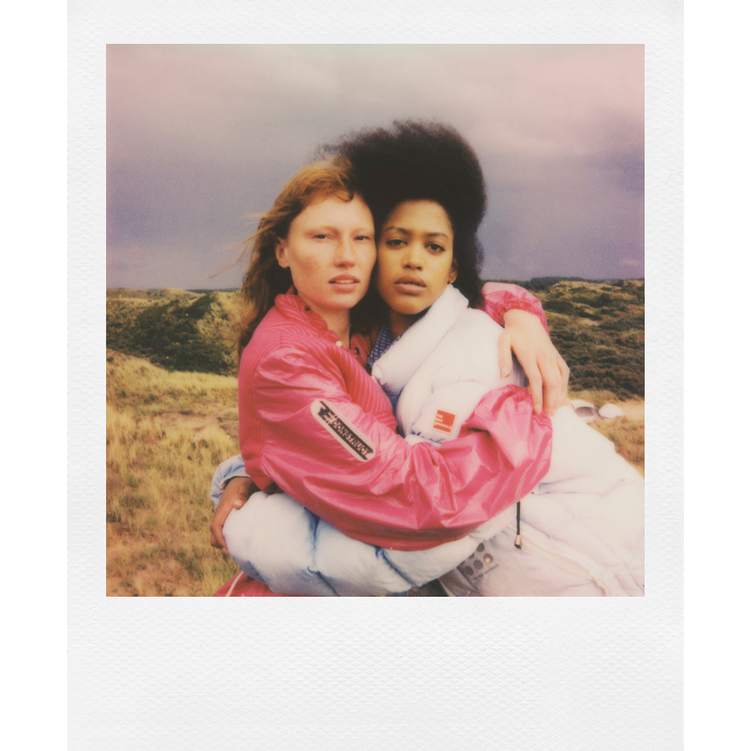 Due donne si abbracciano in una foto Polaroid, su sfondo di un campo erboso e cielo nuvoloso.