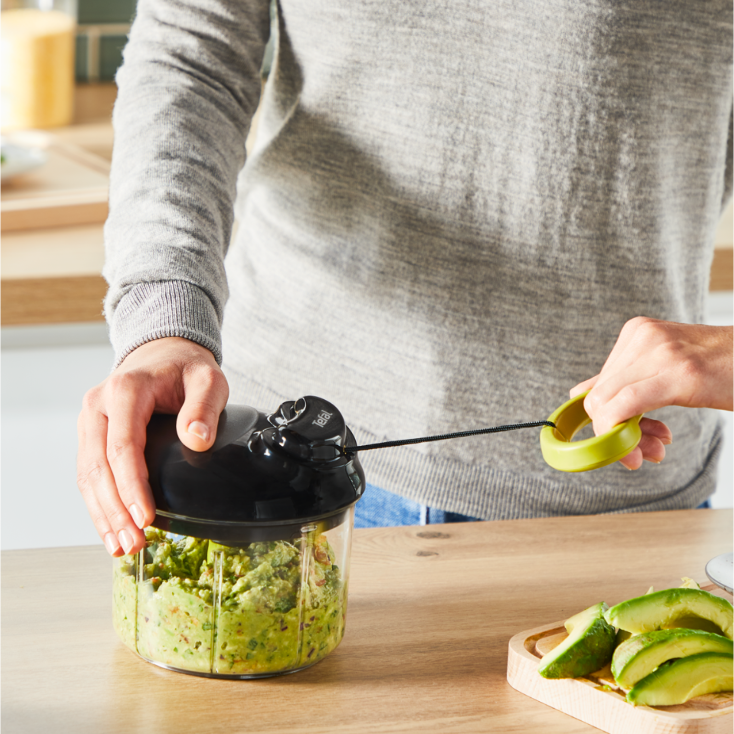 A person chopping avocado in a black Tefal chopper. Avocado slices on a wooden board. White background.