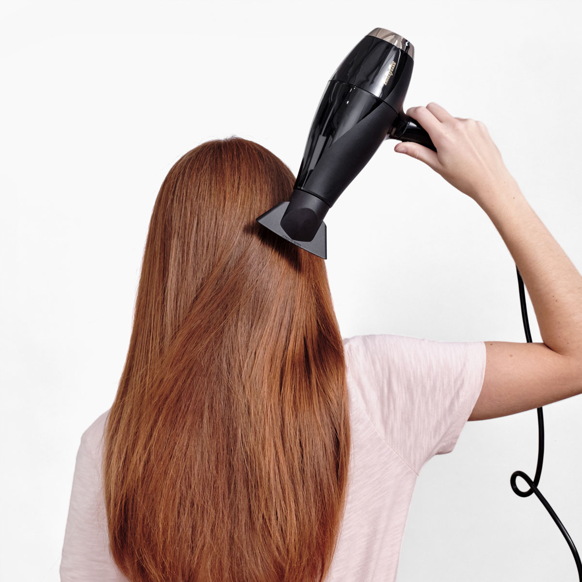 Persona che asciuga lunghi capelli rossi con un asciugacapelli nero, vista da dietro.