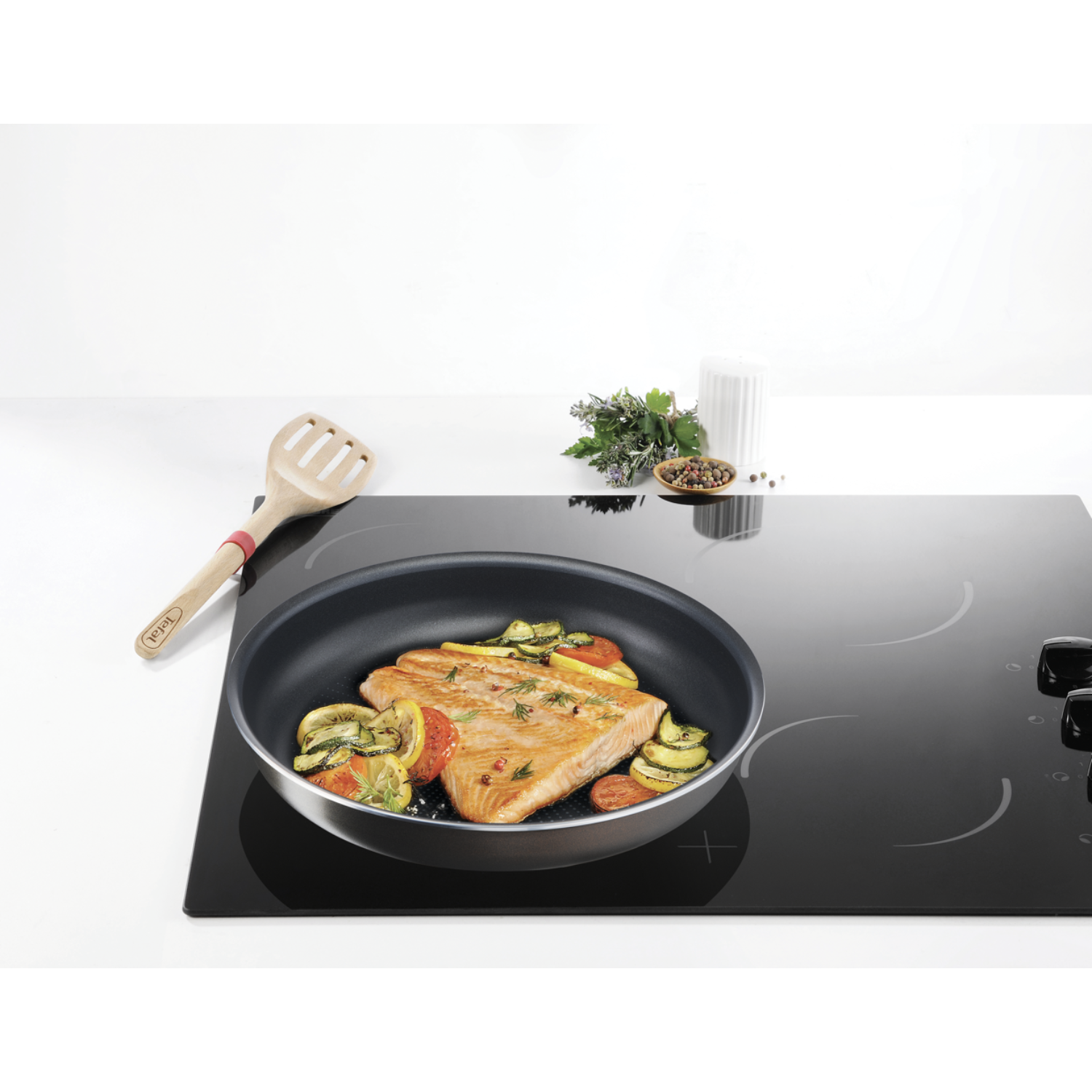 Scène de cuisine : Filet de saumon dans une poêle, avec des légumes, sur une plaque de cuisson noire.