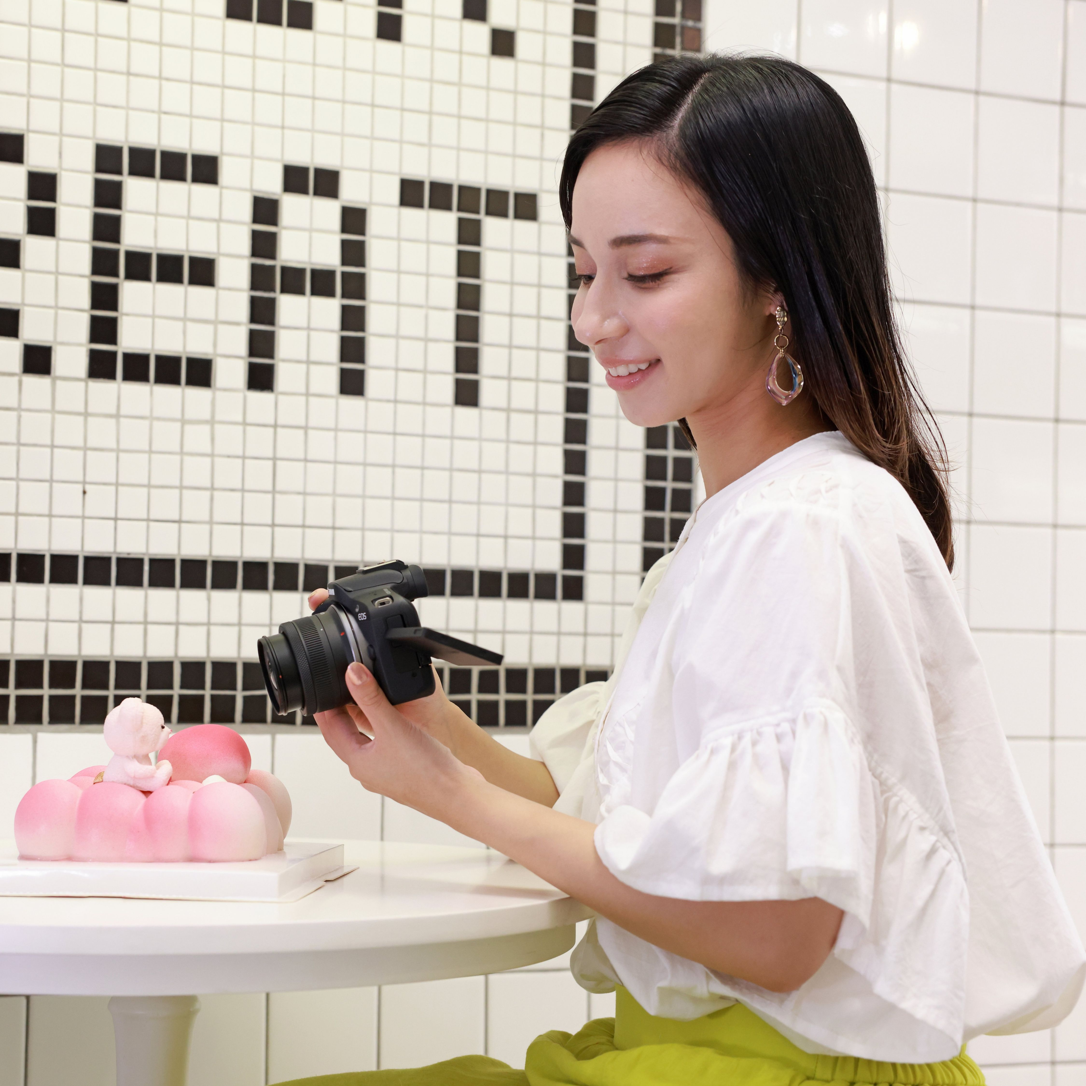 Une femme sourit en tenant un appareil photo, regardant un dessert rose et blanc sur une table.