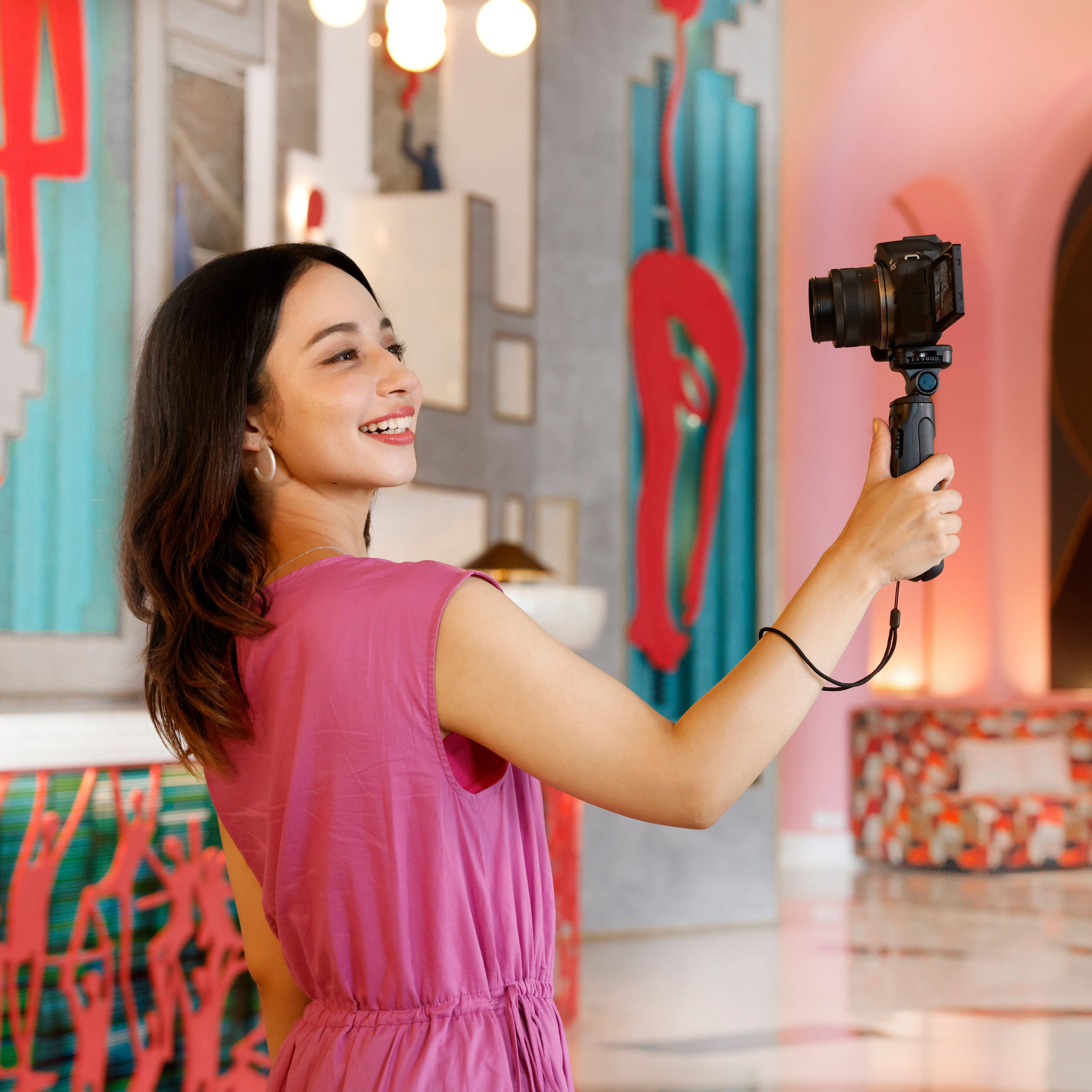 Femme sourit, tenant une caméra sur une perche à selfie. Robe rose, arrière-plan clair.
